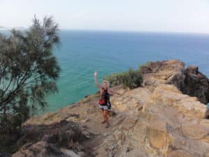 Eine Frau, die auf einem Felsen steht und in Richtung der Kamera winkt, mit Meer im Hintergrund.