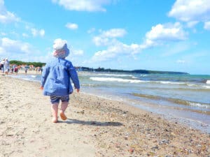 Ein kleines Kind in blauer Jacke und Sonnenhut, das am Strand entlangläuft, mit Wellen im Hintergrund.