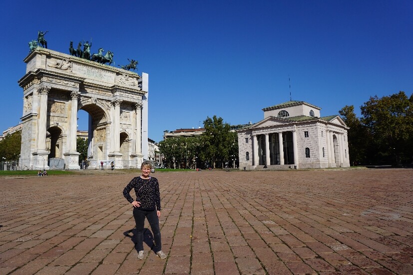 Eine Person steht vor dem Arco della Pace, einem monumentalen Tor in Mailand, umgeben von einem klaren blauen Himmel.
