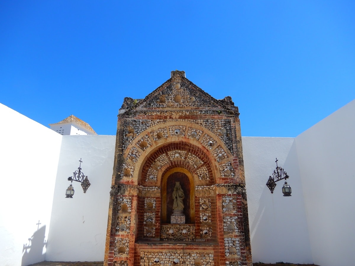 Eine steinerne Nischenfassade mit Statue steht vor weißen Mauern unter klarem blauem Himmel in Faro.