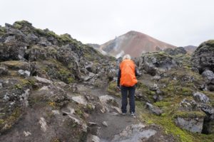 Eine Person in orangefarbener Jacke wandert über felsiges, vulkanisches Terrain mit Rauch aus dem Hintergrund.