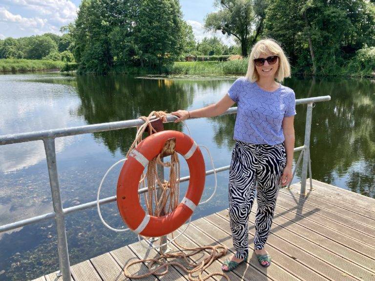 Eine Frau steht neben einem orangefarbenen Schwimmring an einem Steg am Wasser.