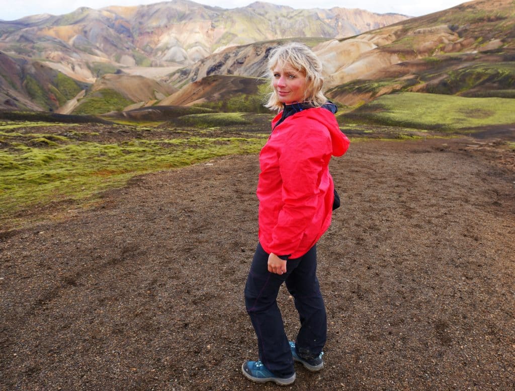 Eine Frau in einer roten Jacke steht in einer bergigen Landschaft mit bunten Hügeln und grünem Gras.