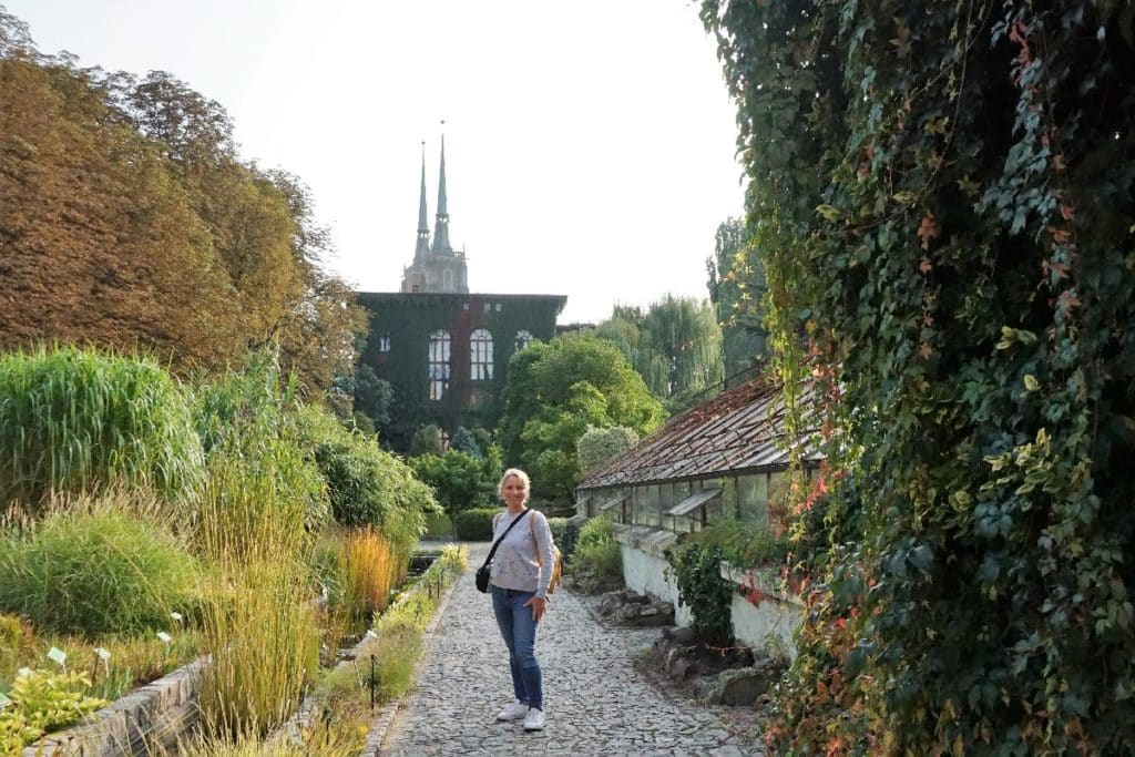 Eine Frau steht in einem Garten mit einem historischen Gebäude und zwei Türmen im Hintergrund in Breslau.