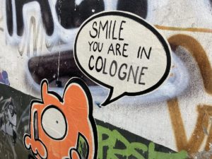 Graffiti mit der Aufschrift 'SMILE YOU ARE IN COLOGNE' und einer orangefarbenen Figur auf einer Wand.