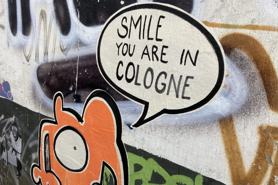 Graffiti mit der Aufschrift 'SMILE YOU ARE IN COLOGNE' und einer orangefarbenen Figur auf einer Wand.