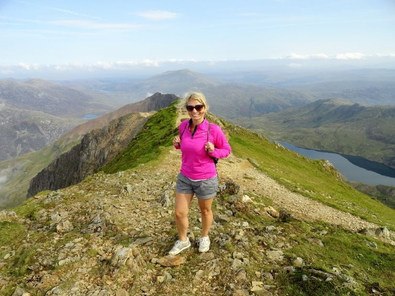 Eine Frau in pinkem Oberteil und Shorts steht auf einem Berggipfel mit Blick auf die Landschaft.