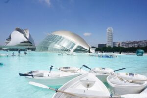 Mehrere kleine Boote im Wasser mit moderner Architektur im Hintergrund, darunter ein gewölbtes Gebäude.
