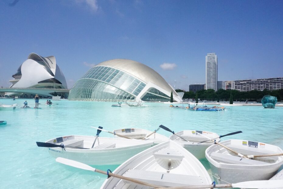 Mehrere kleine Boote im Wasser mit moderner Architektur im Hintergrund, darunter ein gewölbtes Gebäude.