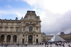 Das Louvre Museum in Paris mit der charakteristischen Glaspyramide im Vordergrund und Besuchern auf dem Platz.