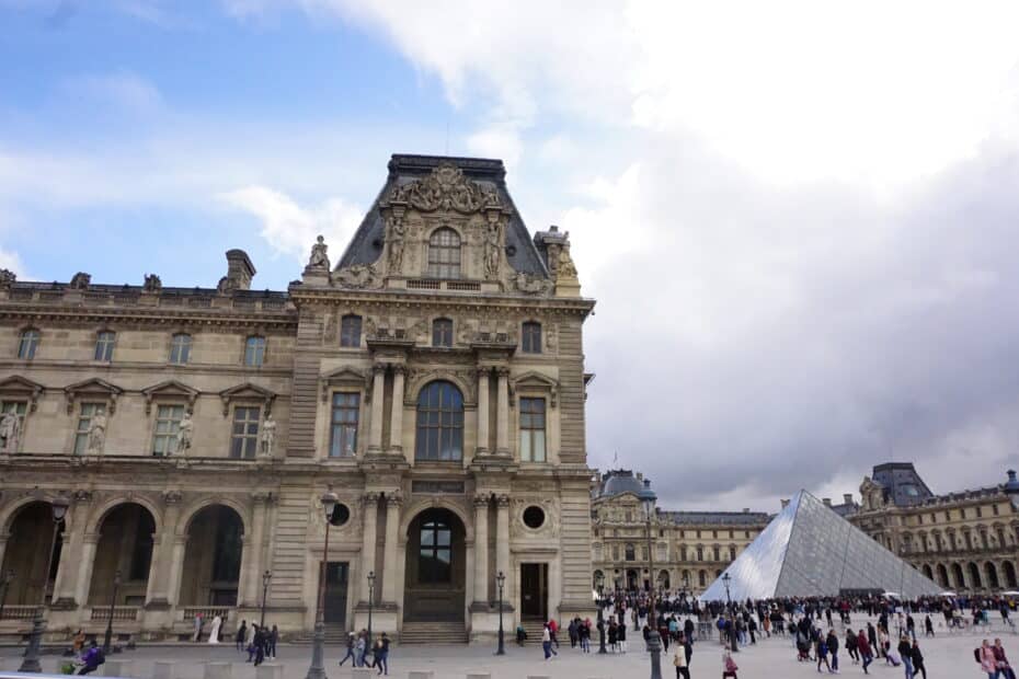 Das Louvre Museum in Paris mit der charakteristischen Glaspyramide im Vordergrund und Besuchern auf dem Platz.