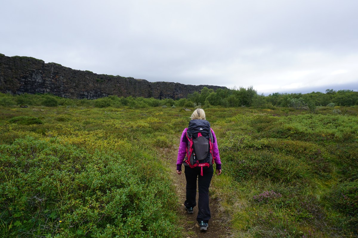 Frau mit Rucksack geht auf einem Pfad durch eine grüne Landschaft mit Felsen im Hintergrund.
