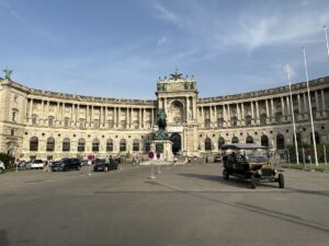 Die Wiener Hofburg mit einem Denkmal und historischen Fahrzeugen im Vordergrund auf einem Platz.