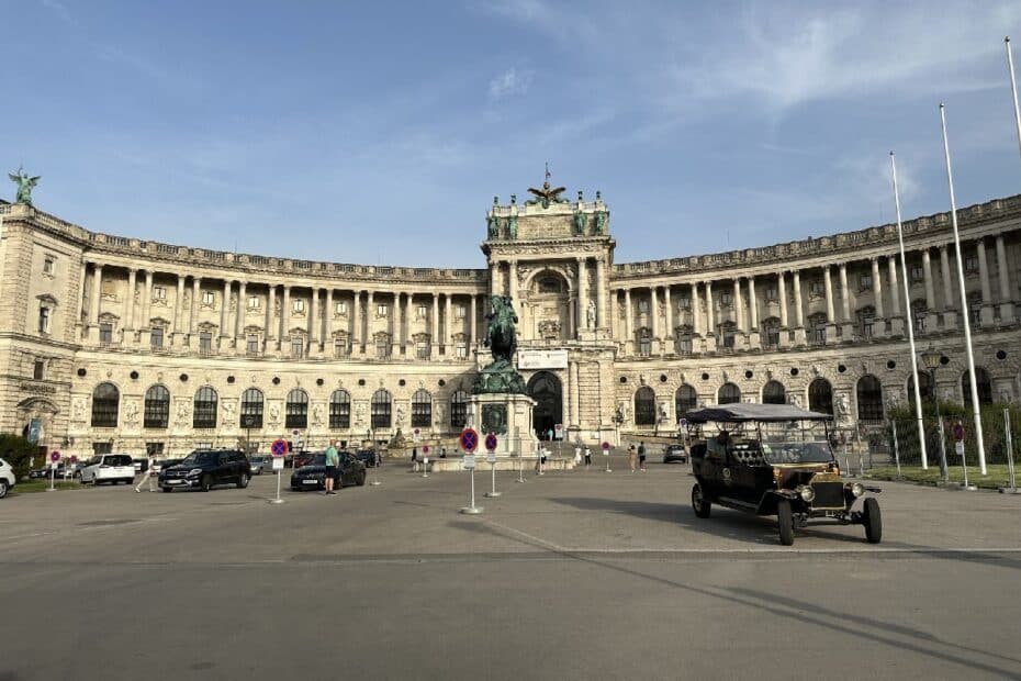 Die Wiener Hofburg mit einem Denkmal und historischen Fahrzeugen im Vordergrund auf einem Platz.