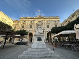Statue auf einem Sockel vor einem historischen Gebäude mit einer Flagge auf dem Dach in Valletta.