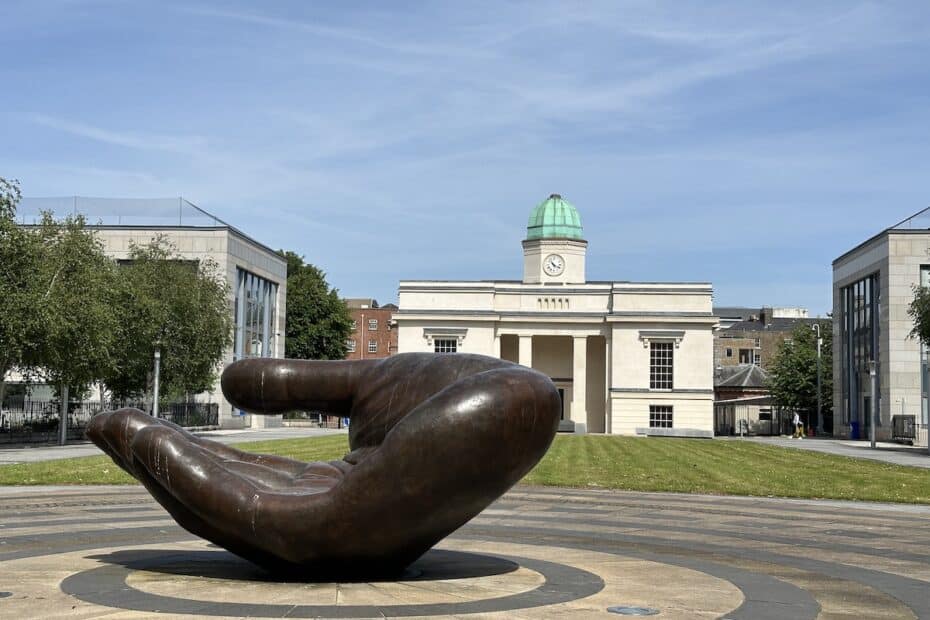 Bronzeskulptur einer großen Hand auf einem Platz in einer städtischen Umgebung.