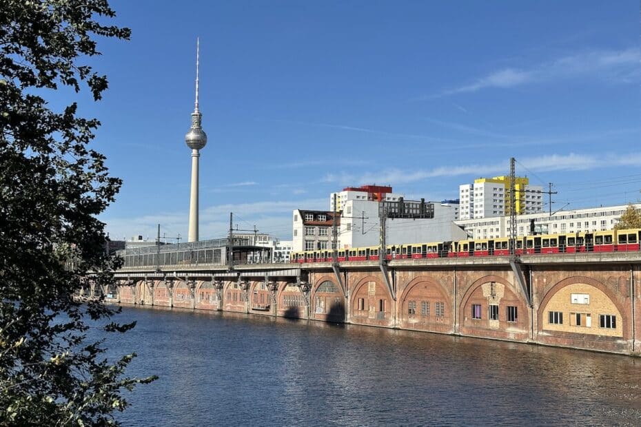 Fernsehturm in Berlin mit einer S-Bahn, die entlang der Spree fährt, im Vordergrund Wasser und Ufervegetation.