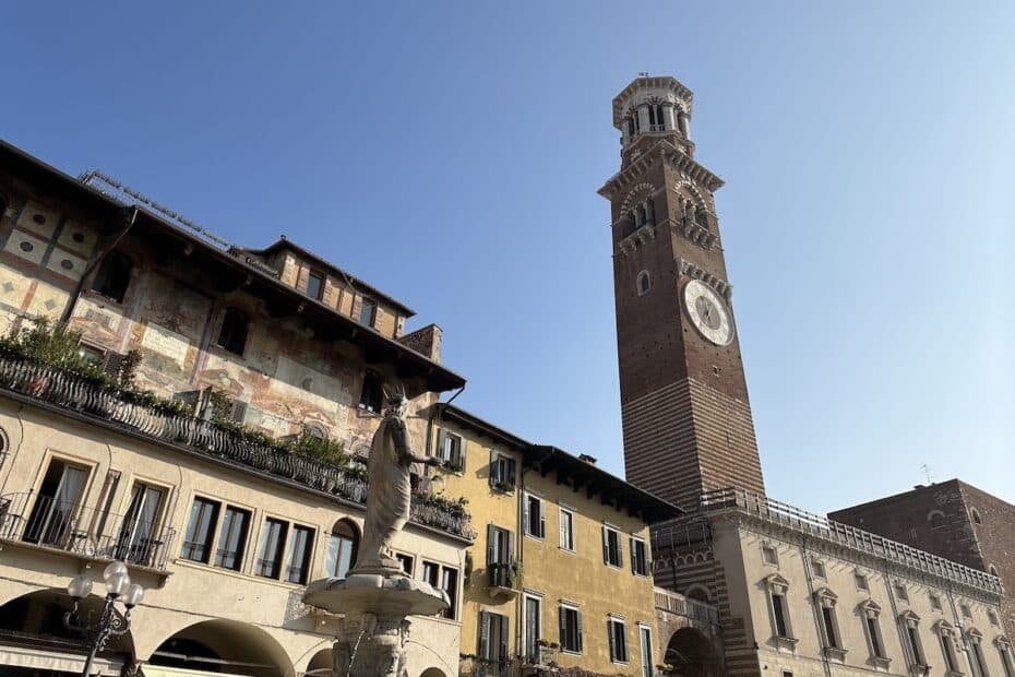 Ein hoher Turm mit einer Uhr und einer Statue im Vordergrund in Verona, Italien, unter klarem Himmel.