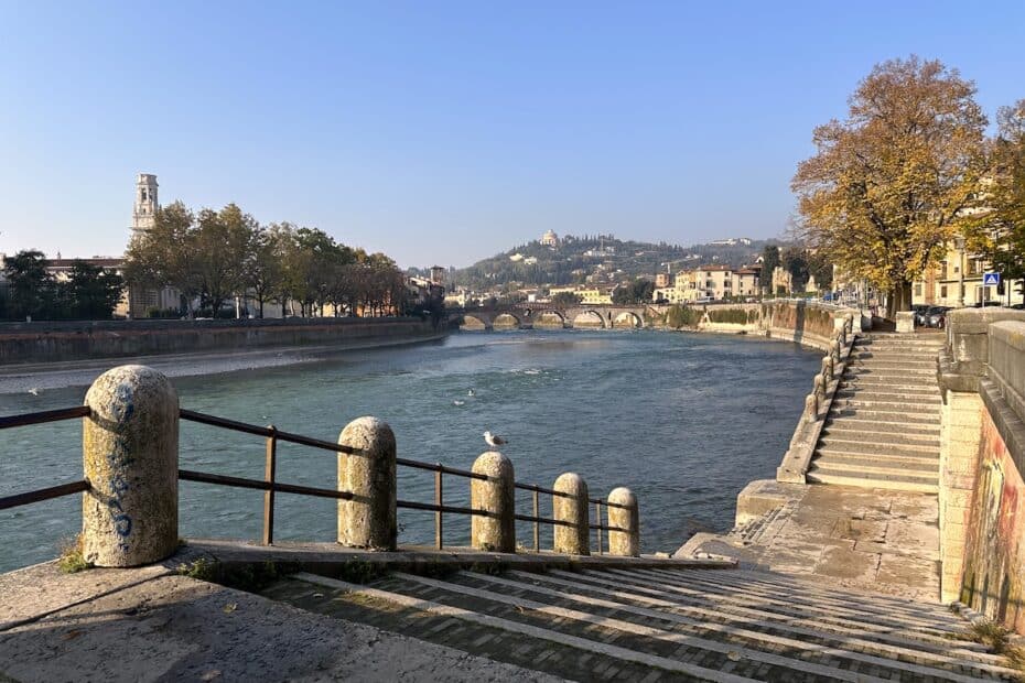 Blick auf einen Fluss mit einer Treppe und einer Brücke in Verona, umgeben von Bäumen und Gebäuden.
