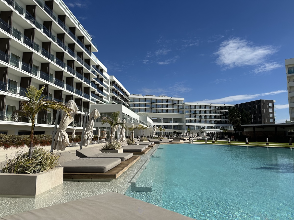 Weitläufiger Pool vor einer modernen Hotelanlage auf Zypern mit Sonnenschirmen, Liegen und türkisfarbenem Wasser