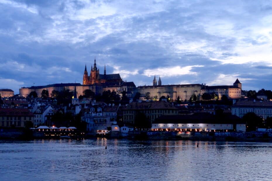Prager Burg und Stadtansicht bei Dämmerung mit reflektierendem Wasser im Vordergrund.