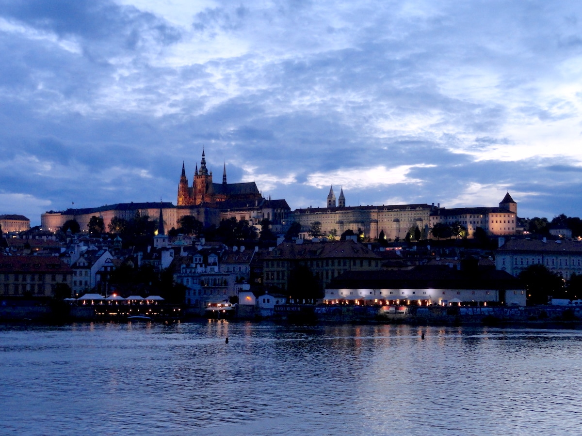 Prager Burg und Stadtansicht bei Dämmerung mit reflektierendem Wasser im Vordergrund.