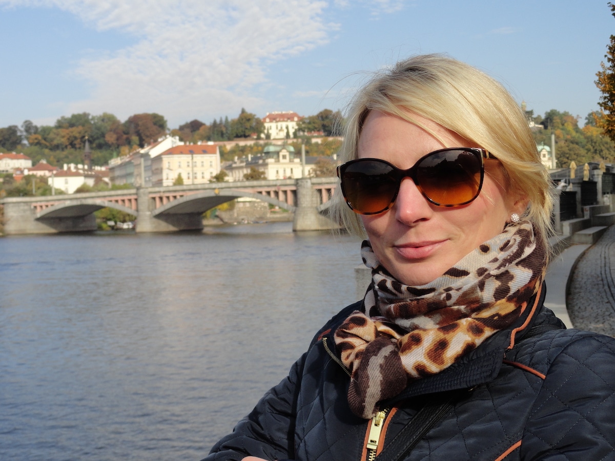 Eine Frau mit blonden Haaren und Sonnenbrille steht am Flussufer mit einer Stadt im Hintergrund.