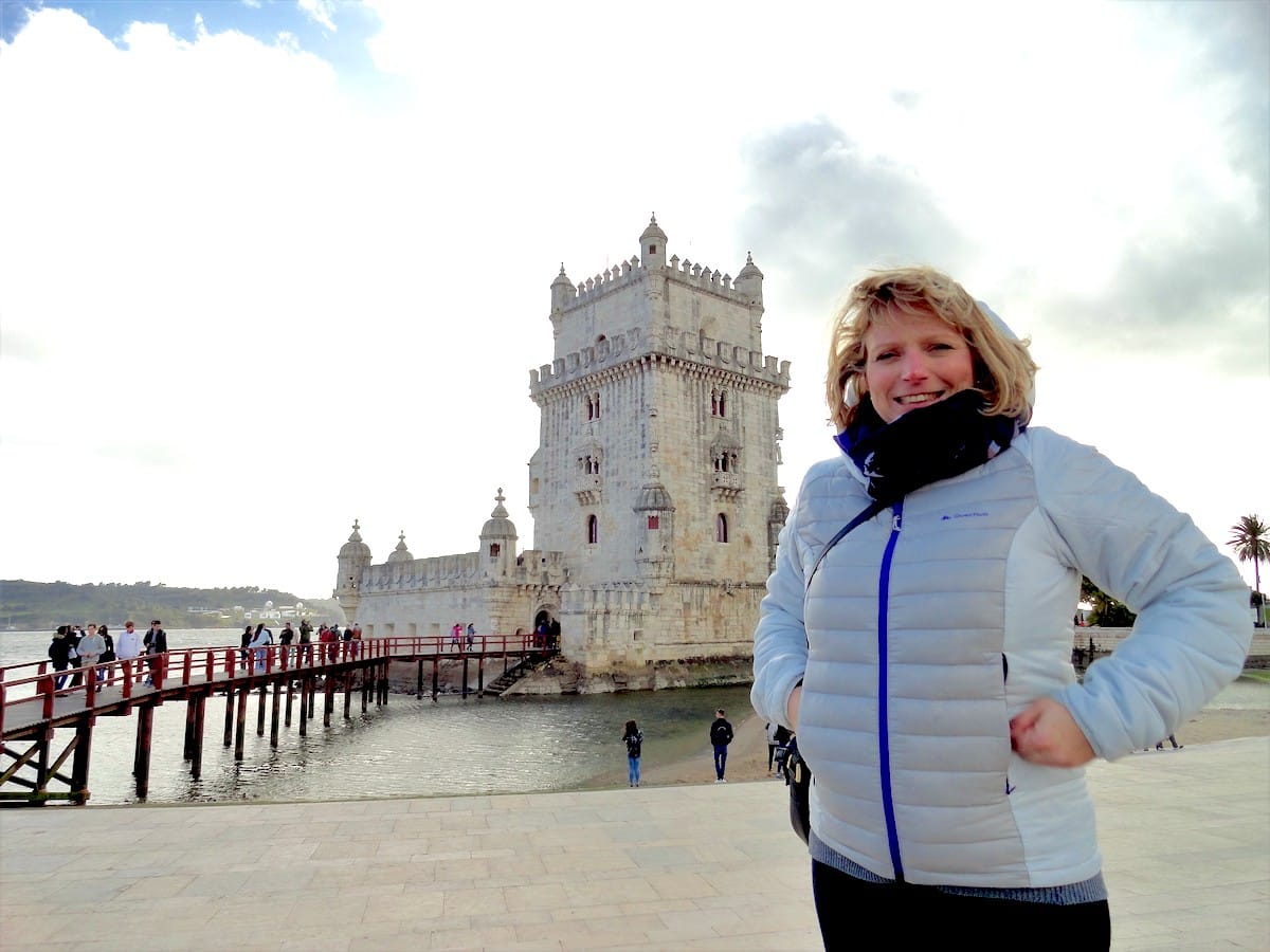 Frau in grauer Jacke steht vor dem Torre de Belém in Lissabon, Portugal, mit Menschen im Hintergrund.