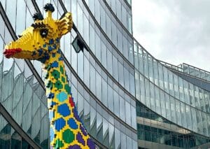 Bunte LEGO-Giraffe, die vor einer modernen Glasfassade steht, mit verschiedenen Farben und Mustern.