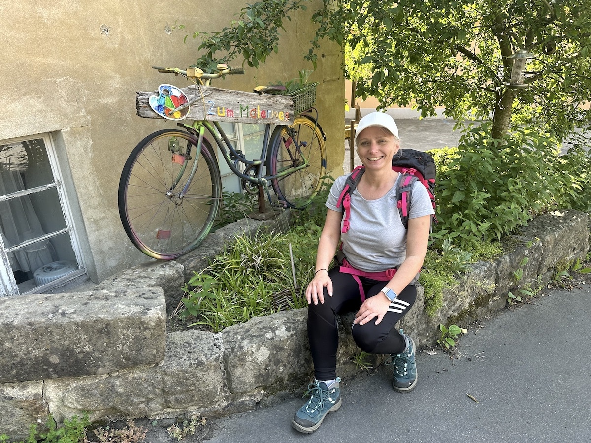 Eine Frau mit einem Rucksack sitzt neben einem bunten Fahrrad, das an einer Wand lehnt.