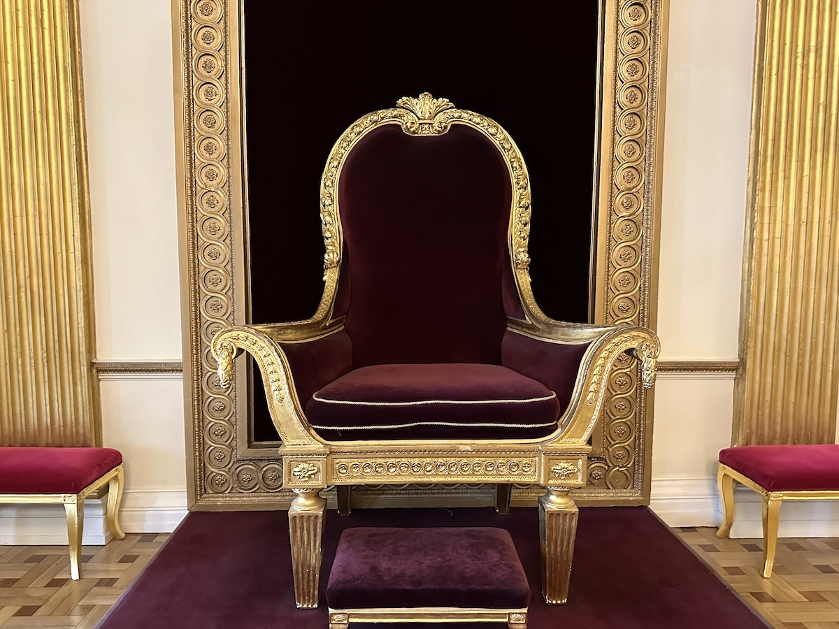 Ein prunkvoller Thron mit goldenen Verzierungen und rotem Samtbezug in einem eleganten Raum.
