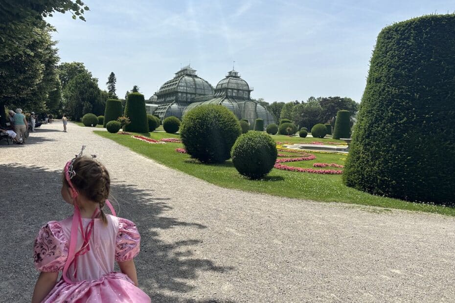 Ein Mädchen in einem rosa Kleid mit einer Krone, das auf einen Garten mit Blumen und Gewächshäusern blickt.