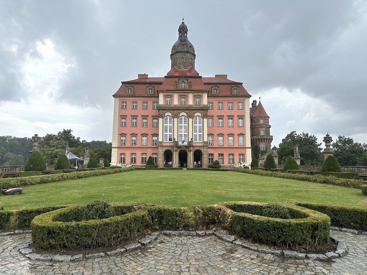 Historisches Schloss mit rosa Fassade und gepflegtem Garten im Vordergrund.