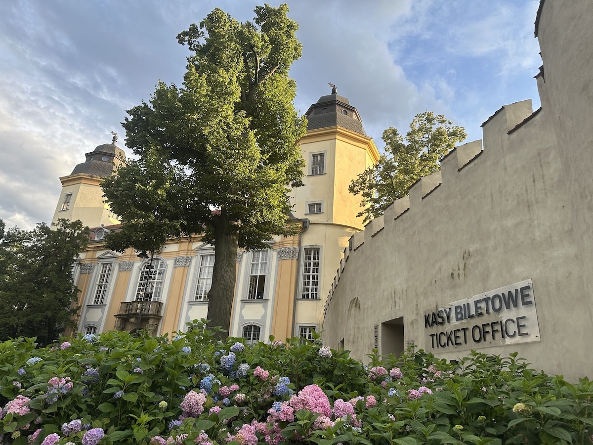 Ticketbüro mit der Aufschrift 'Kasy Biletowe' und historischen Gebäuden im Hintergrund, umgeben von blühenden Pflanzen.