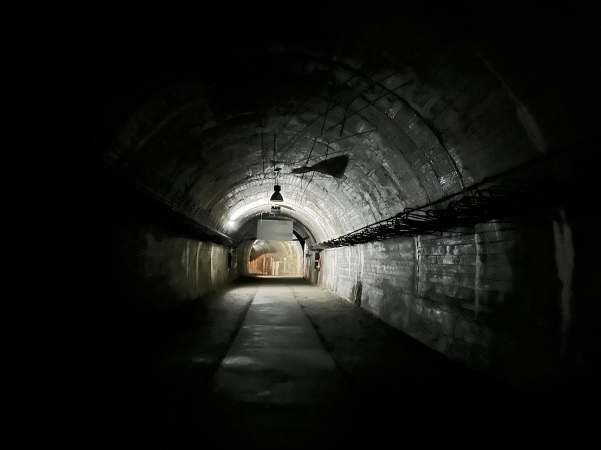 Dunkler Tunnel mit gewölbten Betonwänden und einem beleuchteten Bereich am Ende.