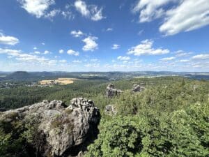 Panoramablick auf eine bewaldete Landschaft mit Felsen und hügeligem Terrain unter einem blauen Himmel mit Wolken.