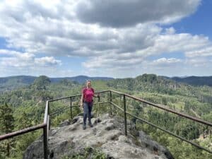Frau in pinkem Shirt steht auf einem Aussichtspunkt mit Blick auf eine bewaldete Landschaft.