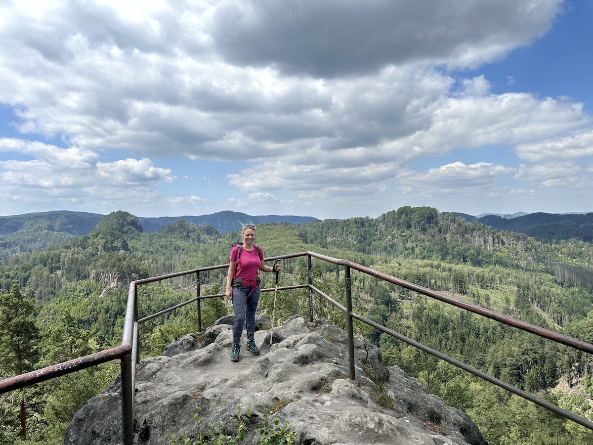 Frau in pinkem Shirt steht auf einem Aussichtspunkt mit Blick auf eine bewaldete Landschaft.