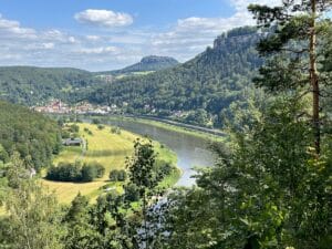 Panoramablick auf eine Flusslandschaft mit bewaldeten Hügeln und Wiesen im Vordergrund.