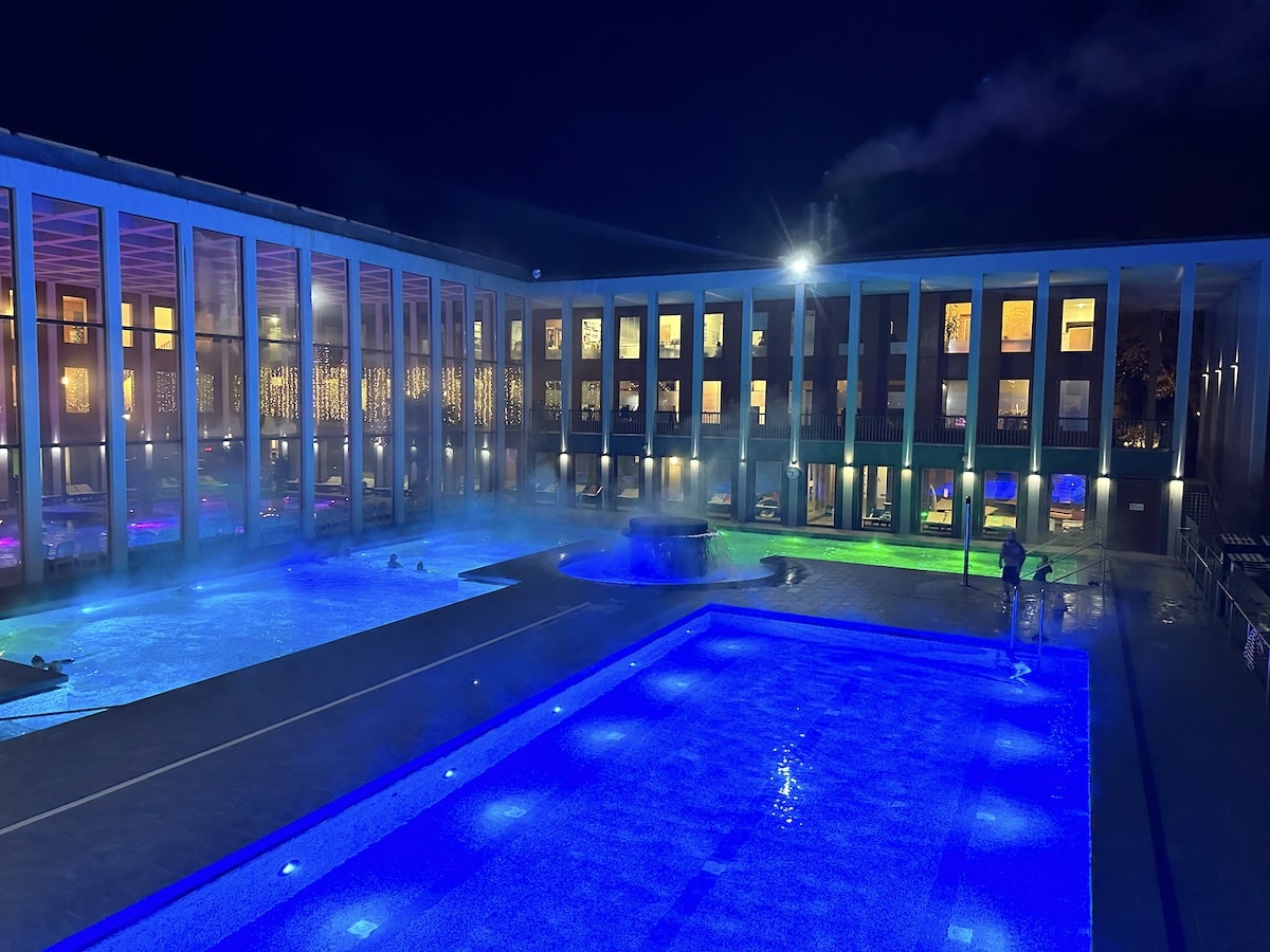 Beleuchteter Außenpool mit Dampfwolken und Wasserfall in einem Hotelkomplex bei Nacht.