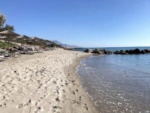 Sandstrand mit Liegen und Sonnenschirmen entlang der Küste und ruhigem Wasser.