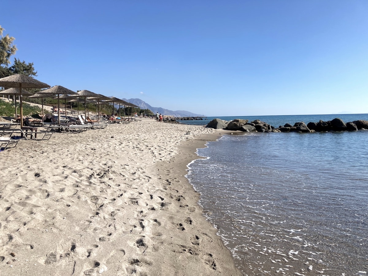 Sandstrand mit Liegen und Sonnenschirmen entlang der Küste und ruhigem Wasser.