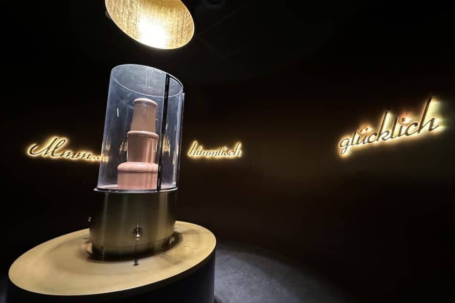 Eine Skulptur in einem Glasbehälter, beleuchtet von oben, mit Schriftzügen an der Wand.