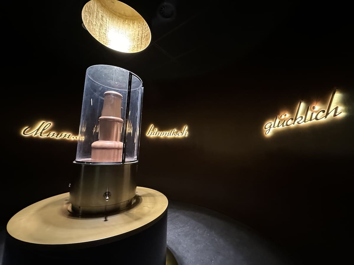 Eine Skulptur in einem Glasbehälter, beleuchtet von oben, mit Schriftzügen an der Wand.