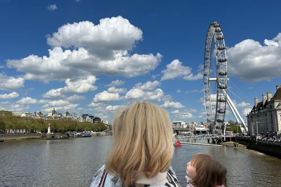 Frau mit blonden Haaren und Kind auf dem Arm, Blick auf das London Eye und den Himmel mit Wolken.