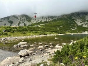 Rote Seilbahn schwebt über einen Bergsee mit Steinen und Grasbewuchs im Vordergrund.