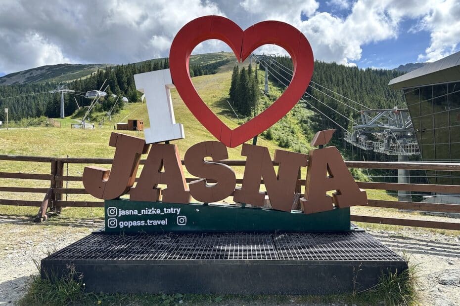 Ein großes, rotes 'I Love Jasná' Schild in den Bergen mit einem Seilbahngebäude im Hintergrund.