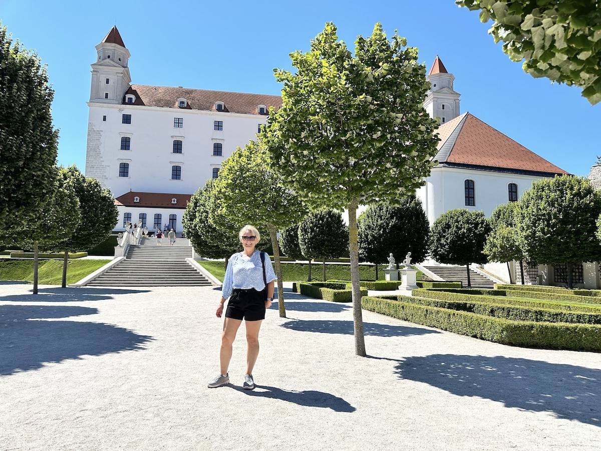 Frau steht im Park mit Bäumen und einem Schloss im Hintergrund an einem sonnigen Tag.