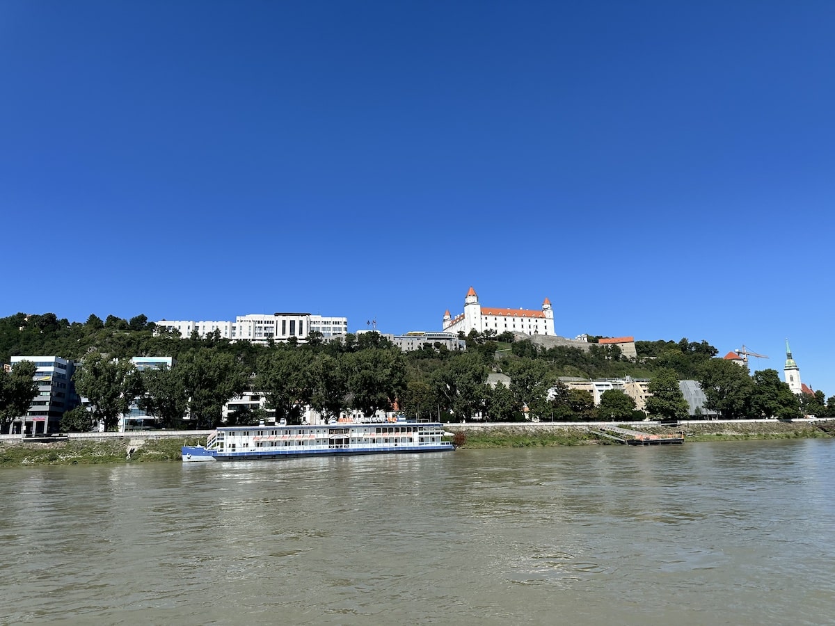 Panoramablick auf die Burg Bratislava mit der Donau im Vordergrund und einem Schiff am Ufer.