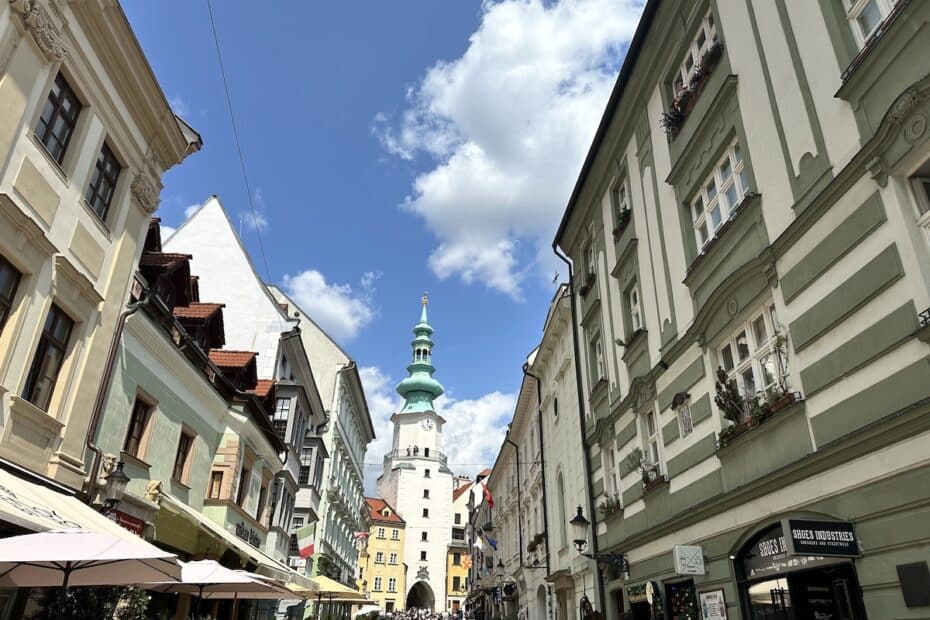 Blick auf eine Straße mit historischen Gebäuden und einem markanten Turm unter einem blauen Himmel mit Wolken.
